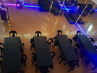 Pride Pole Studio sets up their space using tables with black cloths and chairs in a wooden-floored room with blue and purple neon lights and festive string lights on poles.