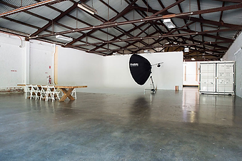 Spacious studio at Fridays Studio with wooden beams, white walls, and a shiny floor. A table with chairs and a large lighting setup are in view.