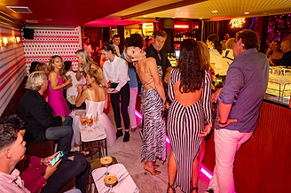 Party at a bar at Sassy Perth with elegantly dressed people. Woman in zebra dress poses playfully. Neon "Aperol Spritz" sign glows. Festive atmosphere.