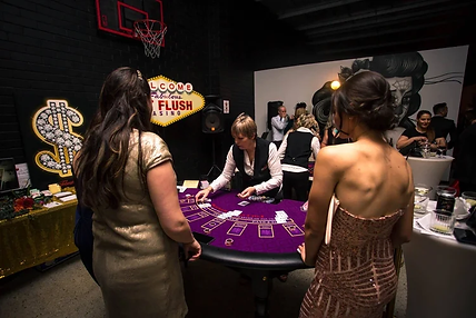 People in elegant attire play a card game at a casino-themed party. A dealer handles cards on a purple table. set up at The Hens House